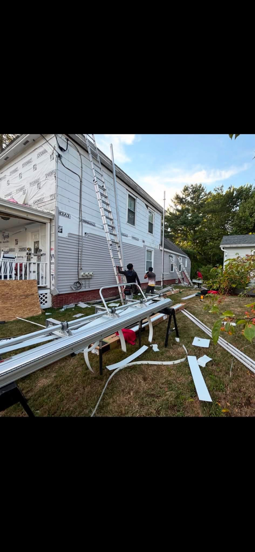 Crew installing new vinyl siding with ladders and gutter stock on site